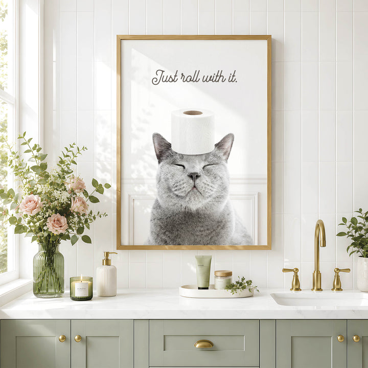 a gray cat with its eyes closed, wearing a roll of toilet paper on its head, and sitting in front of a framed picture on a bathroom wall.