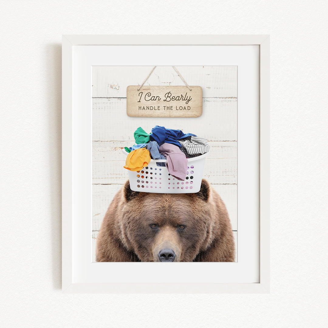 A brown bear sits atop a laundry basket filled with clothes, with a sign above that reads "I Can Bearly Handle the Load".
