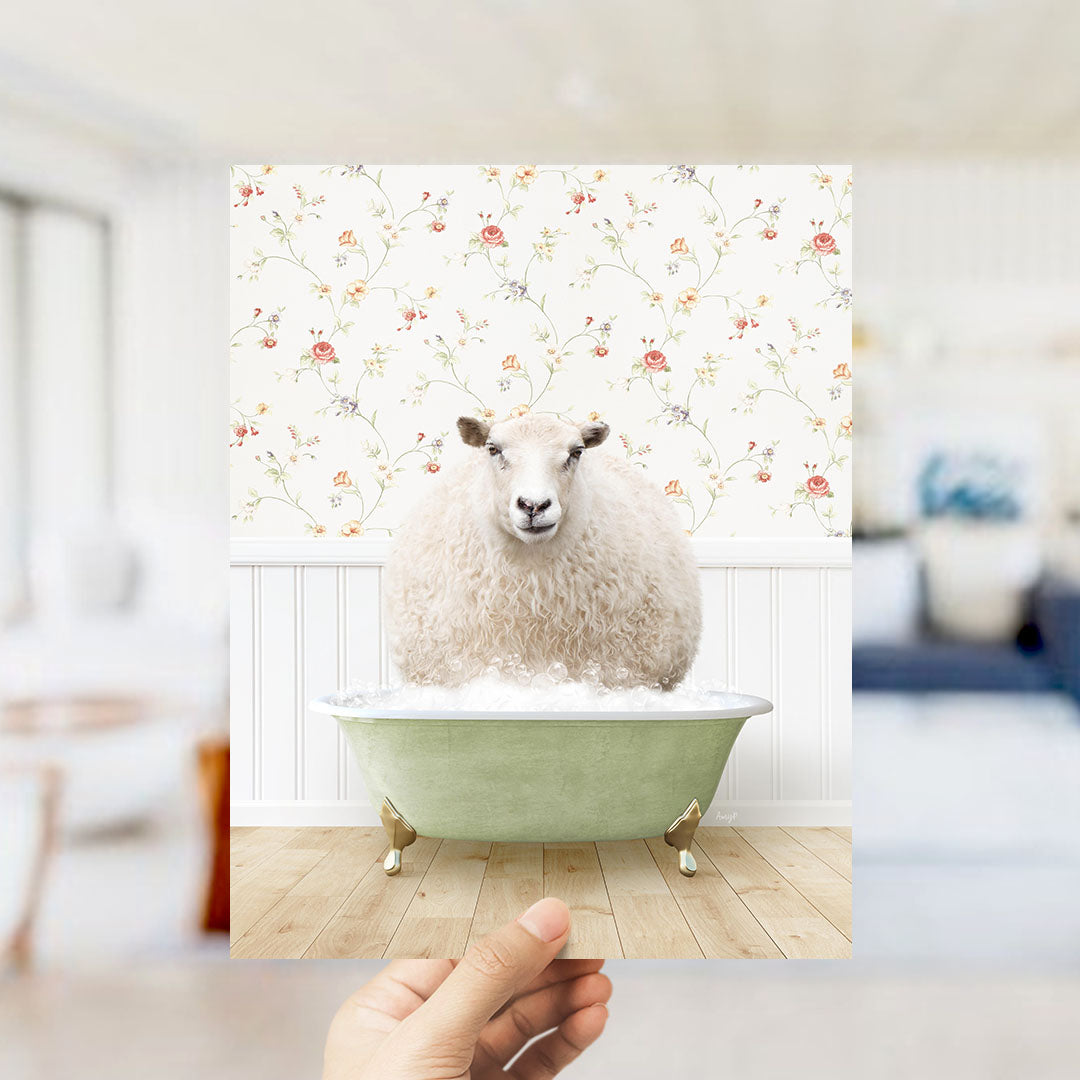 A hand holds up a framed image of a sheep sitting in a green bathtub, with a floral wallpaper in the background.
