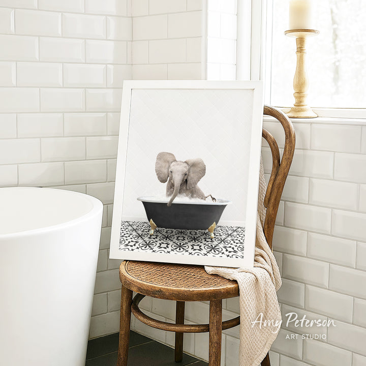 A framed image of an elephant sitting in a bathtub is displayed on a wooden chair in a bathroom setting.