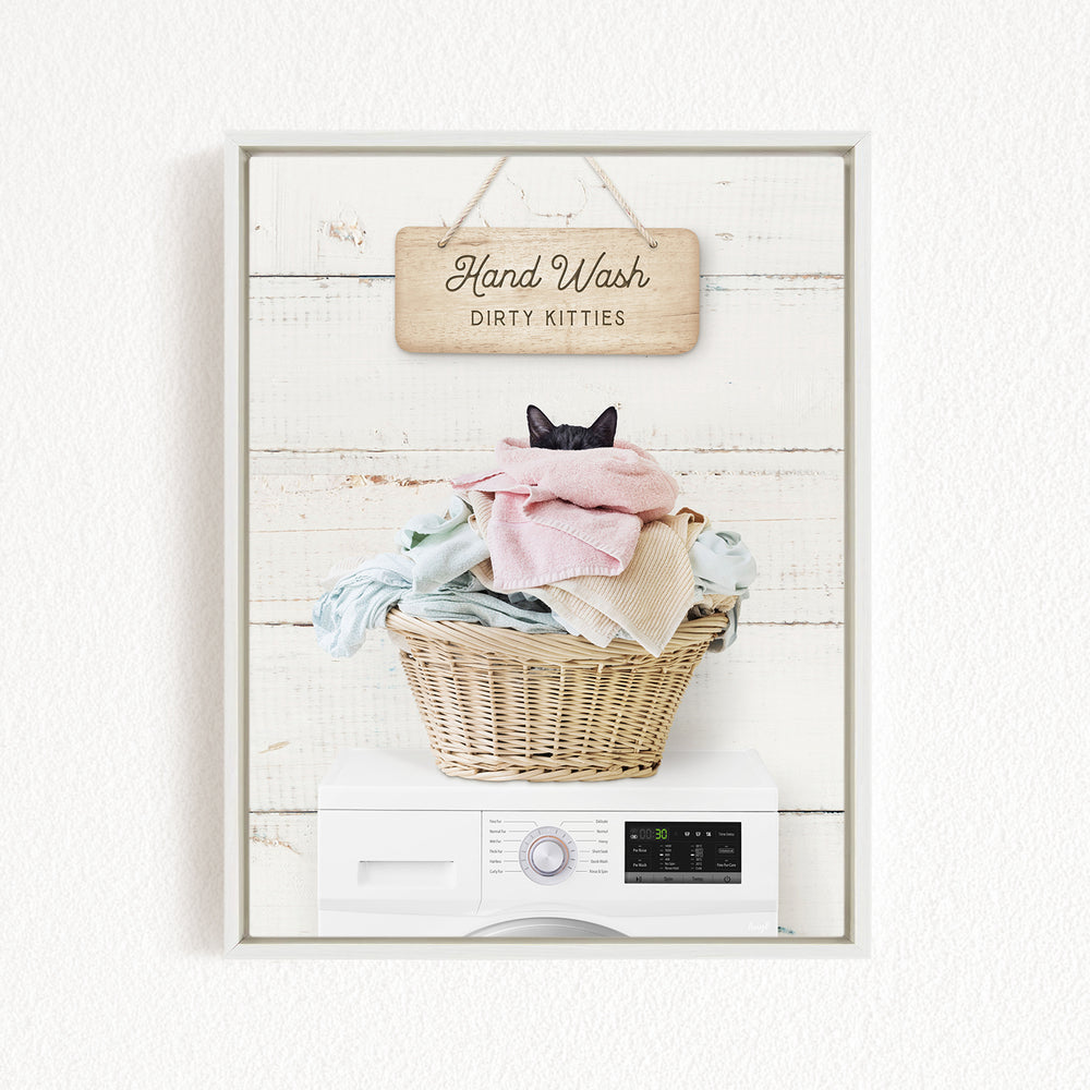 A wooden sign with the text "Hand Wash Dirty Kitties" hangs above a wicker basket filled with laundry, which includes a black cat peeking out from the clothes.