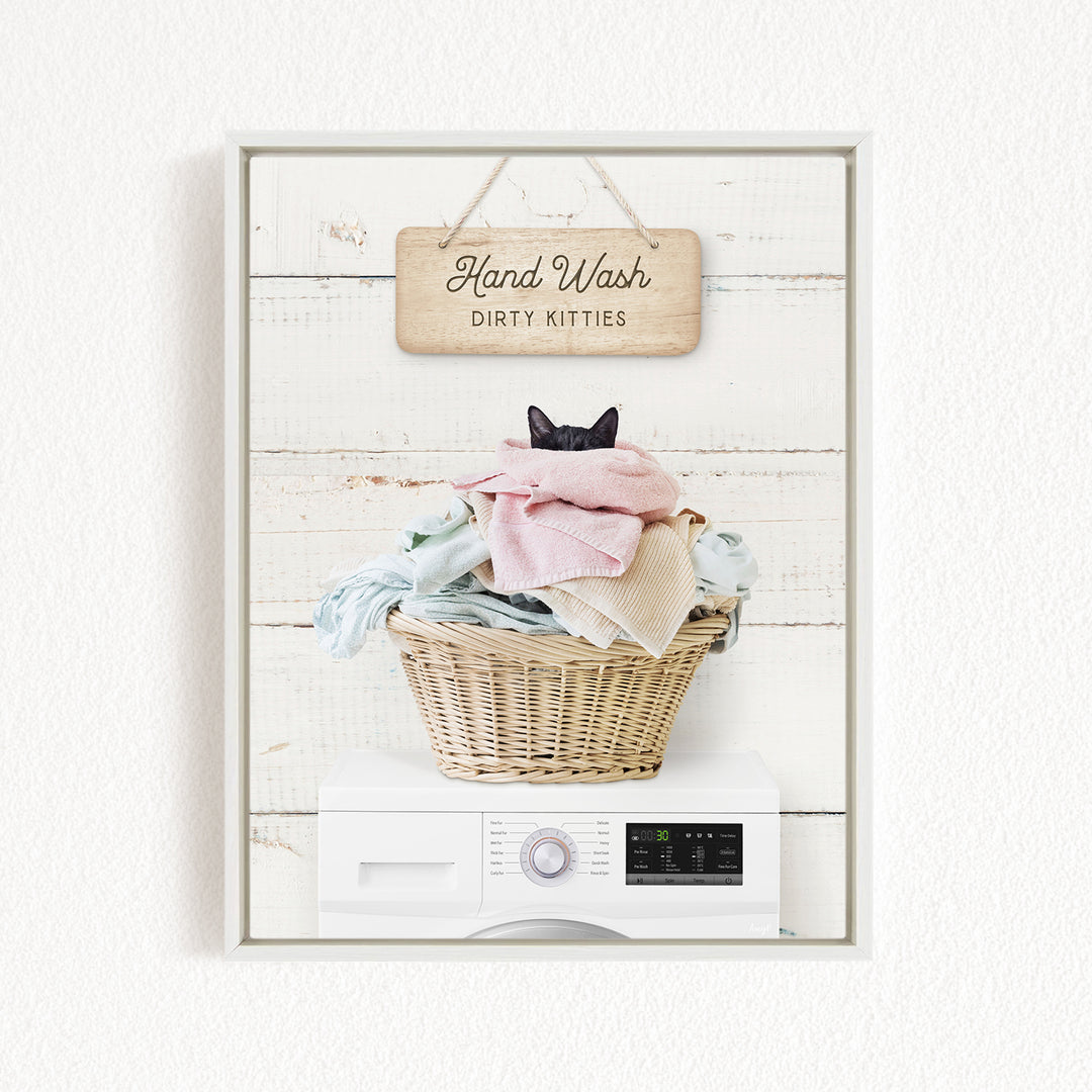 A wooden sign with the text "Hand Wash Dirty Kitties" hangs above a wicker basket filled with laundry, which includes a black cat peeking out from the clothes.