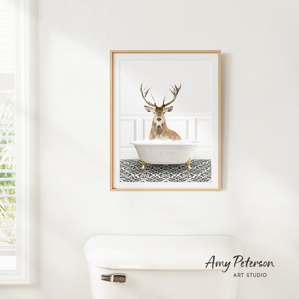A framed image of a deer sitting in a bathtub, with a patterned tile floor beneath it, is hanging on a white wall.