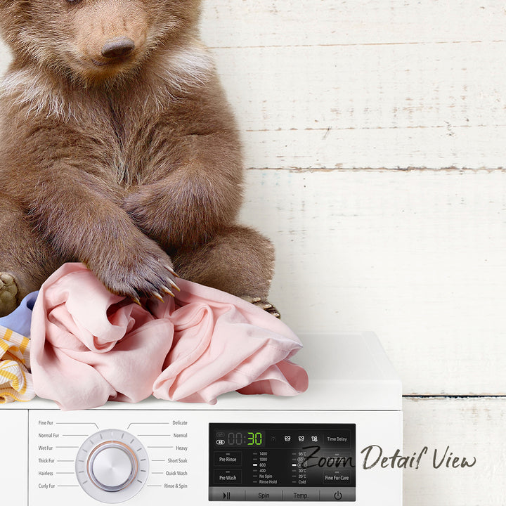 A brown bear sitting on a pile of clothes next to a white washing machine.