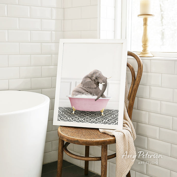A framed photograph of a gray elephant sitting in a pink bathtub, with a patterned rug on the floor.