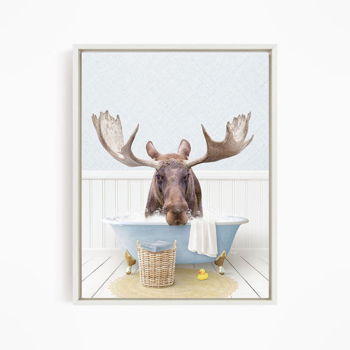 A moose with large antlers is sitting in a blue bathtub filled with water, surrounded by a yellow rubber duck and a basket.