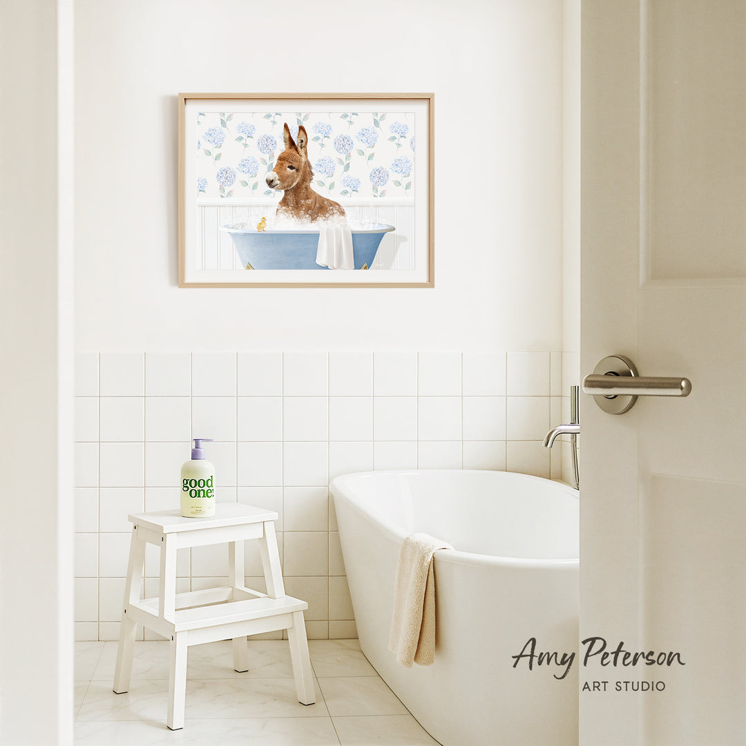 A framed painting of a dog sitting in a bathtub is hanging on a white tiled wall above a white bathtub in a bathroom.
