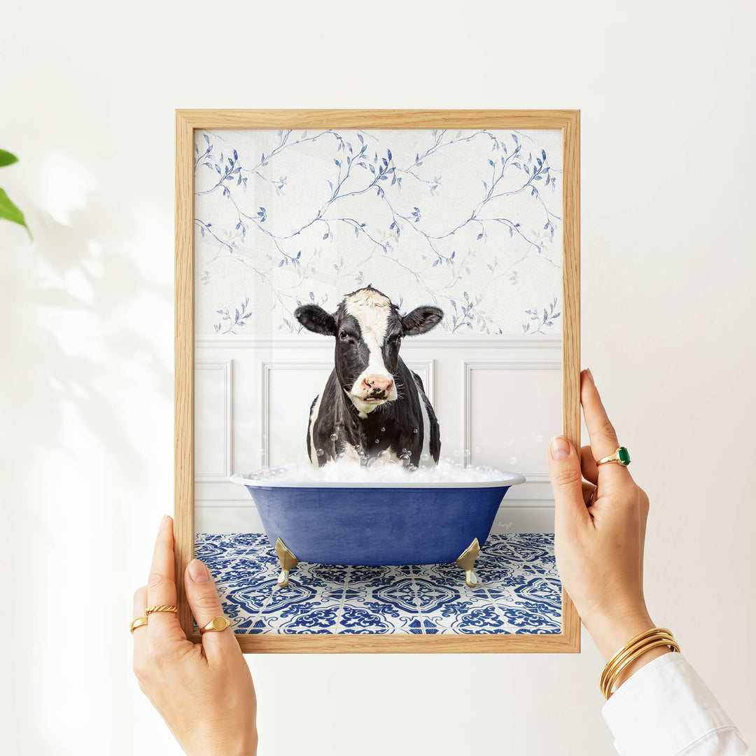 A person's hands are holding a framed picture of a black and white cow sitting in a blue bathtub filled with water, surrounded by a blue and white floral pattern.