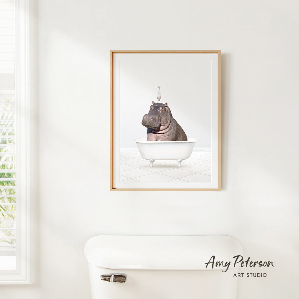 A framed image of a hippopotamus sitting in a bathtub, with a window and a toilet visible in the background.