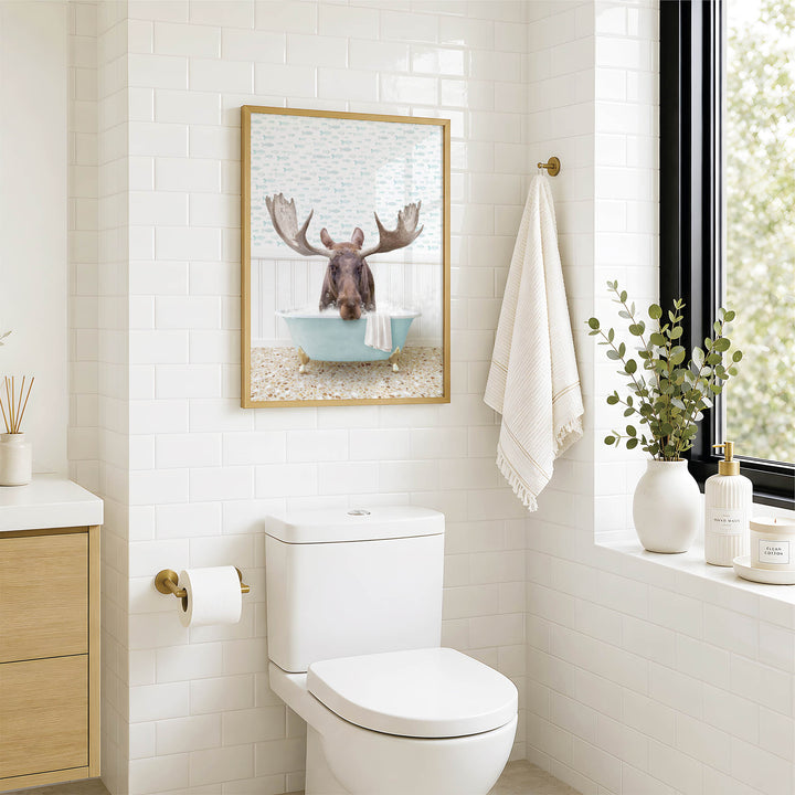 A bathroom with a white toilet, a framed picture of a moose in a bathtub, and a window with greenery on the sill.