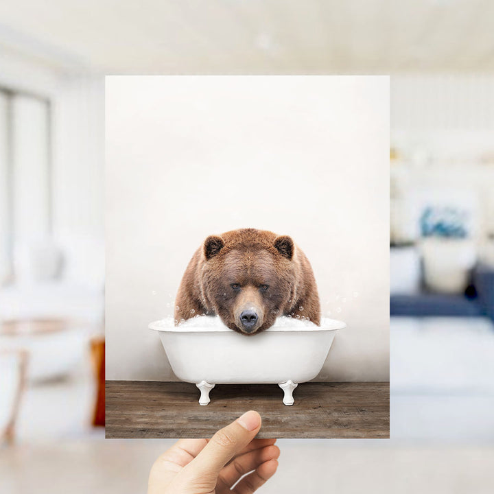 A hand holds up a framed image of a brown bear sitting in a white bathtub filled with water.