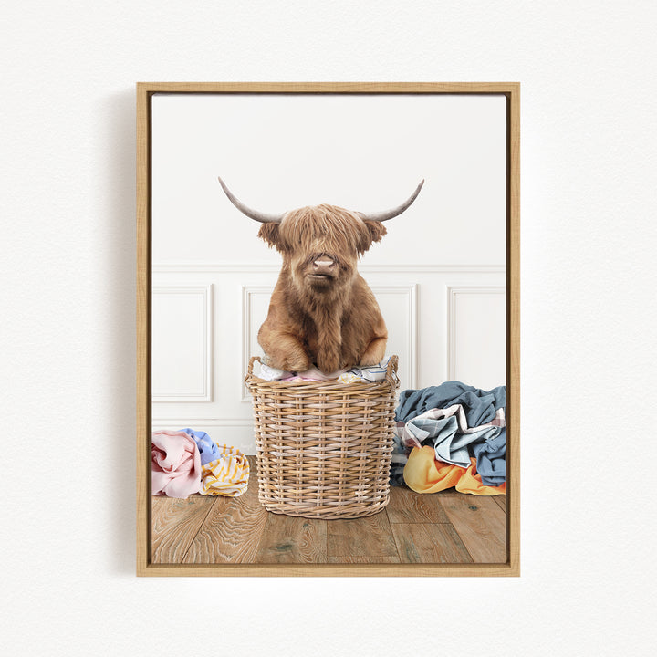 A brown Highland cow with large horns sits in a wicker basket filled with clothes, creating a whimsical and playful scene.