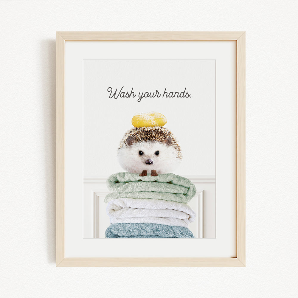 A framed image of a hedgehog sitting atop a stack of folded towels, with the text "Wash your hands" written above it.
