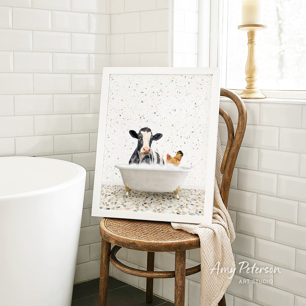 A framed image of a cow and a chicken sitting in a bathtub, with a towel draped over the back of a wooden chair in a bathroom setting.