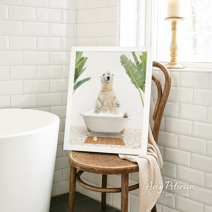 A framed image of a polar bear sitting in a bathtub, surrounded by tropical plants, is displayed on a wooden chair in a bathroom setting.