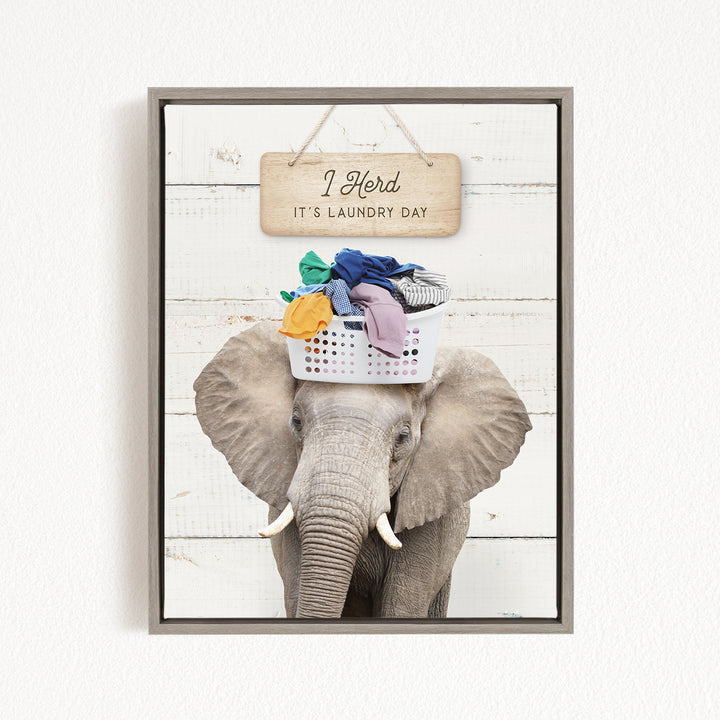 A gray elephant standing in front of a wooden sign that says "It's laundry day" with a basket of clothes on its head.