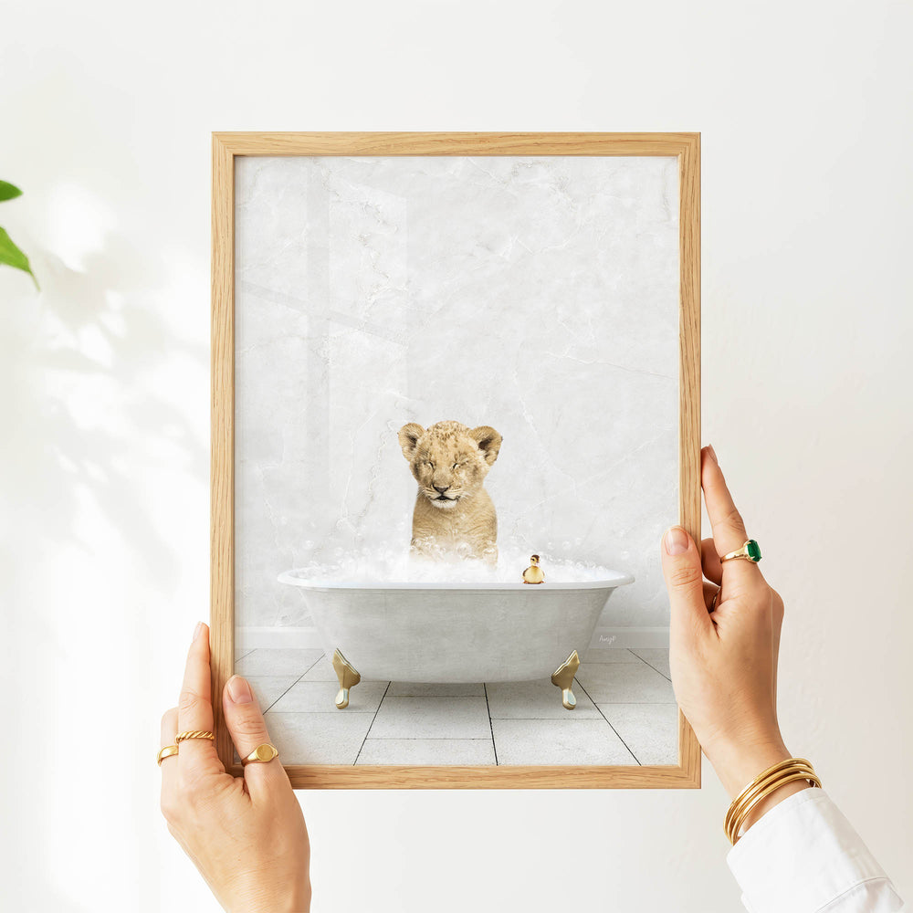 A person's hands are holding a framed picture of a baby lion sitting in a bathtub filled with water and bubbles.