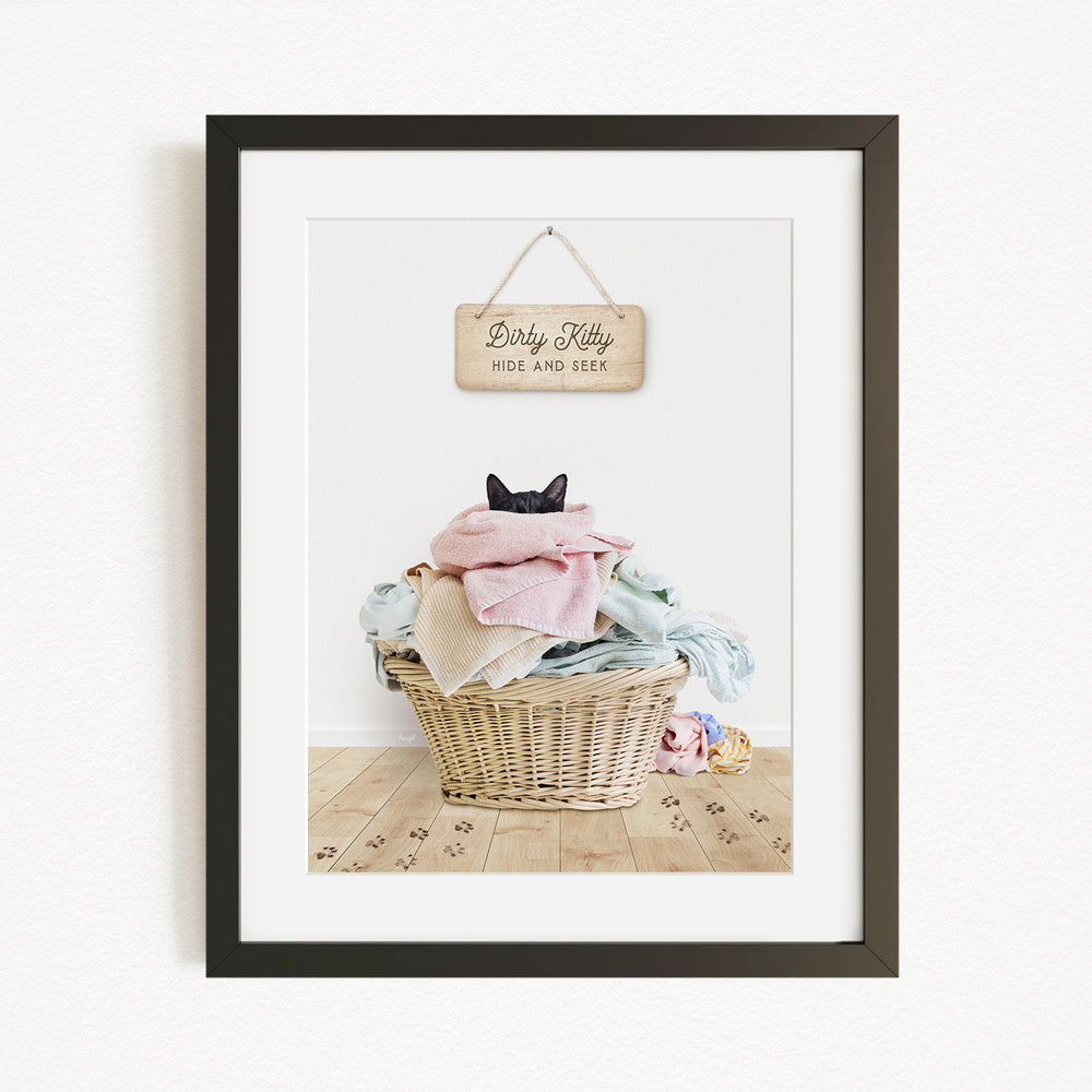 A framed photograph of a cat sitting in a wicker basket filled with clothes, with a sign above the basket that reads "Dirty Kitty HIDE AND SEEK".