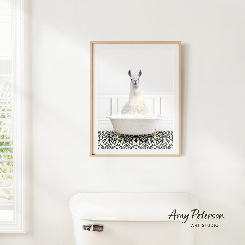 A framed image of a white llama sitting in a bathtub is hanging on a white wall in a bathroom.