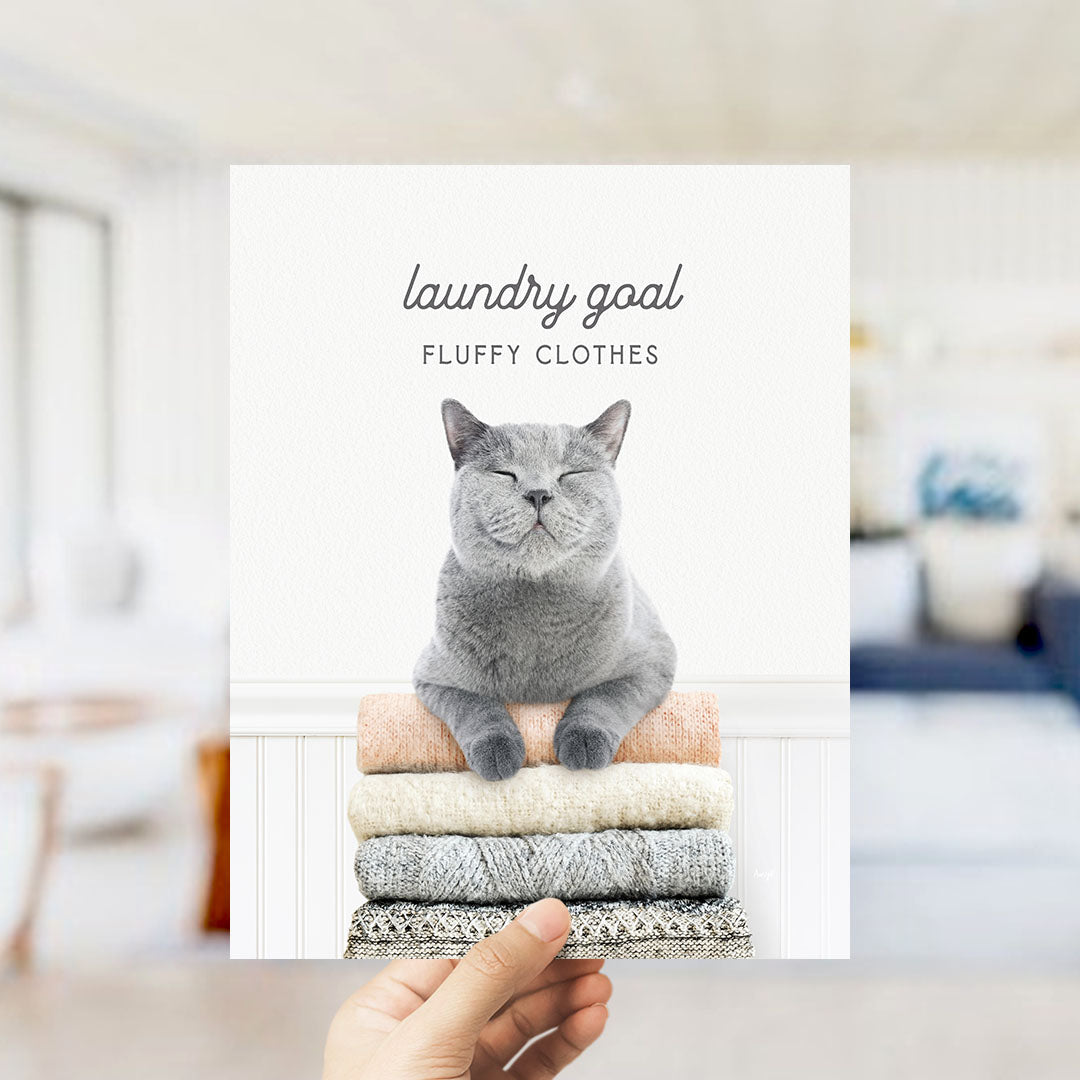 A hand holding a stack of laundry with a smiling gray cat sitting on top of the clothes.