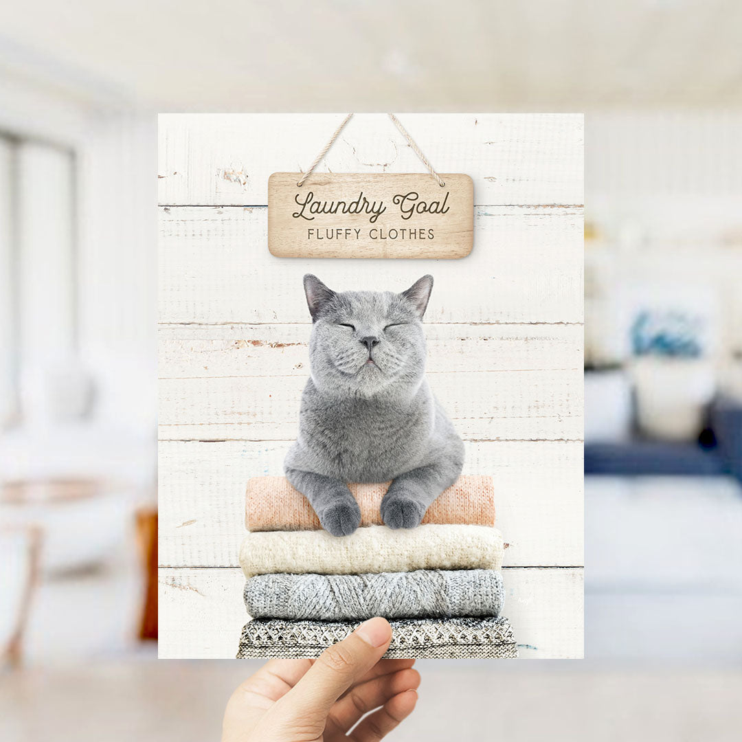 A hand holding a framed image of a gray cat sitting on a stack of laundry, with a sign above that says "Laundry Goal Fluffy Clothes".