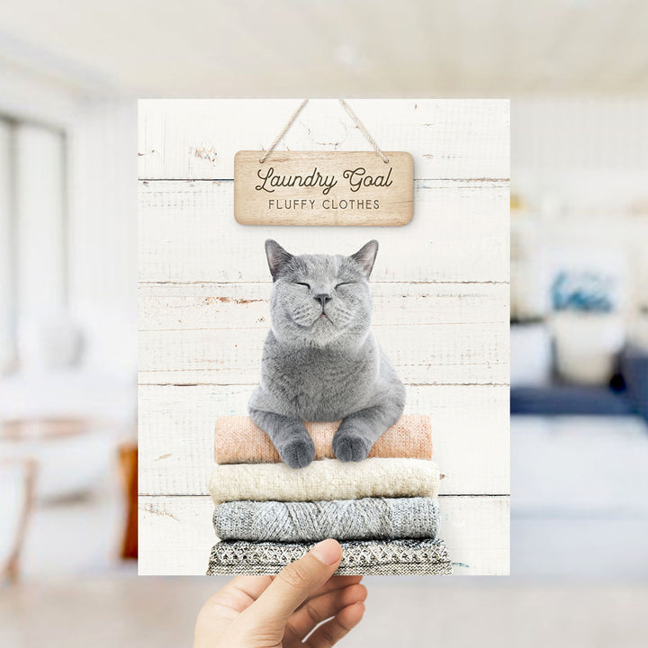 A hand holding a framed image of a gray cat sitting on a stack of laundry, with a sign above that says "Laundry Goal Fluffy Clothes".