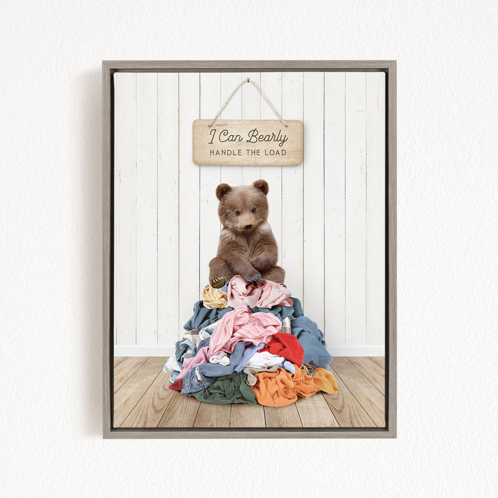 A teddy bear sitting atop a pile of clothes, with a wooden sign above that reads "I Can Bearly Handle the Load".