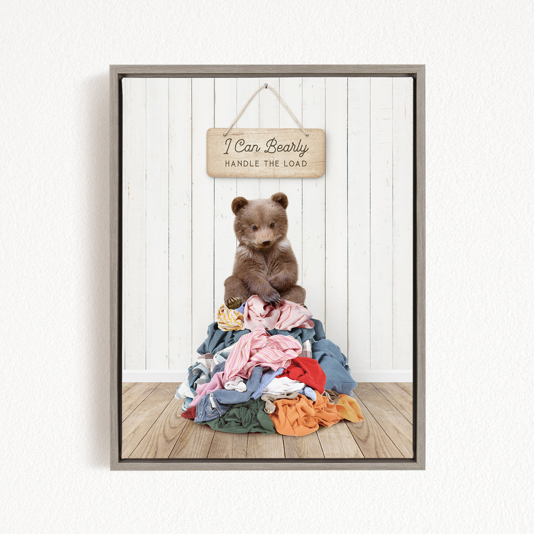 A teddy bear sitting atop a pile of clothes, with a wooden sign above that reads "I Can Bearly Handle the Load".
