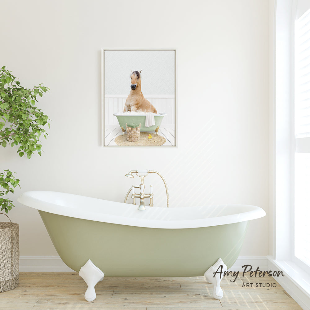 A framed image of a horse sitting in a bathtub is displayed on the wall above a green clawfoot bathtub in a bright, white-walled bathroom.