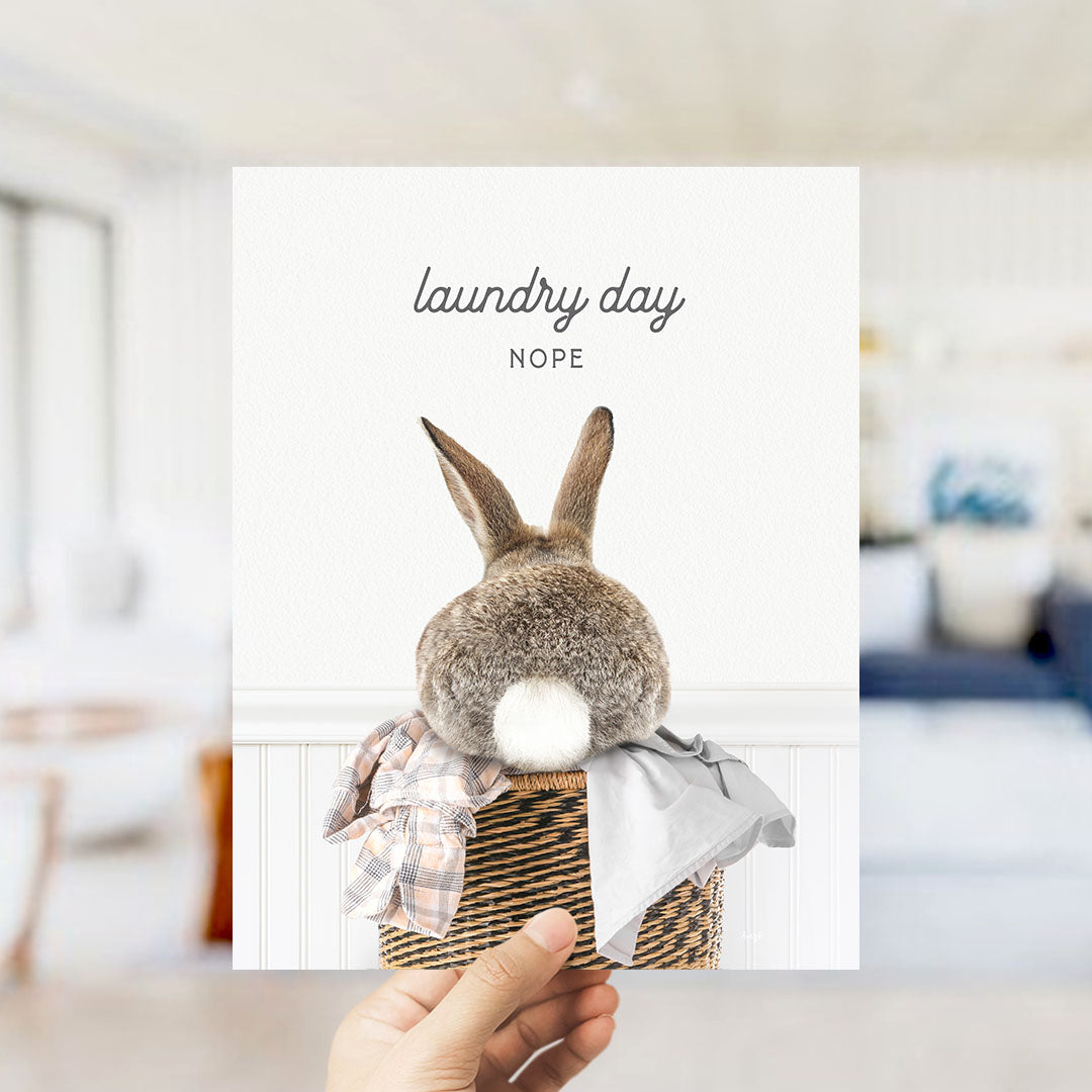 A hand holding a card with a picture of a rabbit sitting in a basket, with the text "laundry day NOPE" written above it.