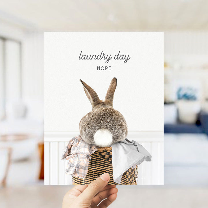 A hand holding a card with a picture of a rabbit sitting in a basket, with the text "laundry day NOPE" written above it.