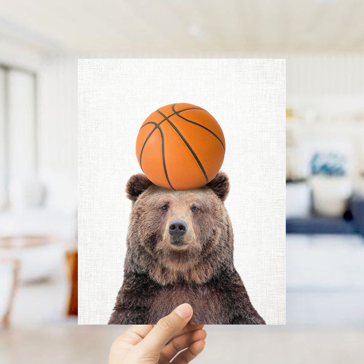 A hand holds up a picture of a brown bear with a basketball on its head.