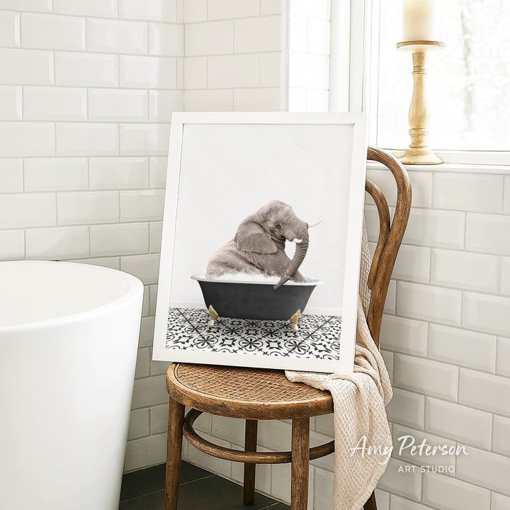 A framed image of a gray elephant sitting in a bathtub, surrounded by a patterned tile floor, is displayed on a wooden chair in a bathroom setting.
