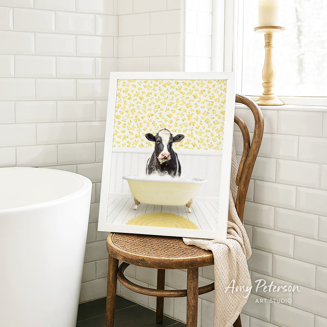 A framed image of a cow sitting in a bathtub, surrounded by yellow flowers, is displayed on a wooden chair in a bathroom setting.