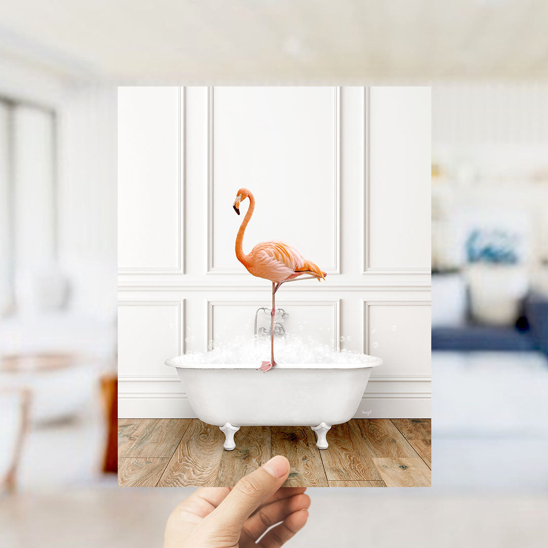 A hand holds up a framed image of a pink flamingo standing on a white bathtub filled with water, creating a whimsical and playful scene.