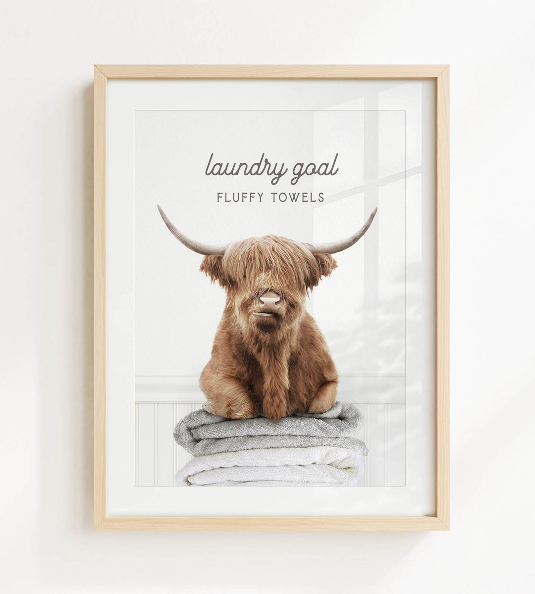 A framed image of a brown cow with long horns sitting on a stack of fluffy towels, with the text "laundry goal" and "fluffy towels" written above it.