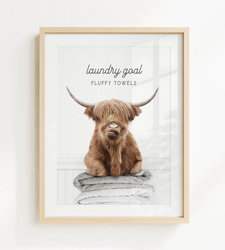 A framed image of a brown cow with long horns sitting on a stack of fluffy towels, with the text "laundry goal" and "fluffy towels" written above it.