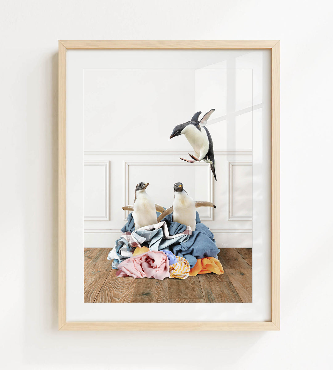 A framed photograph depicting two penguins standing on a pile of clothes, with one penguin flying in the air above the pile.