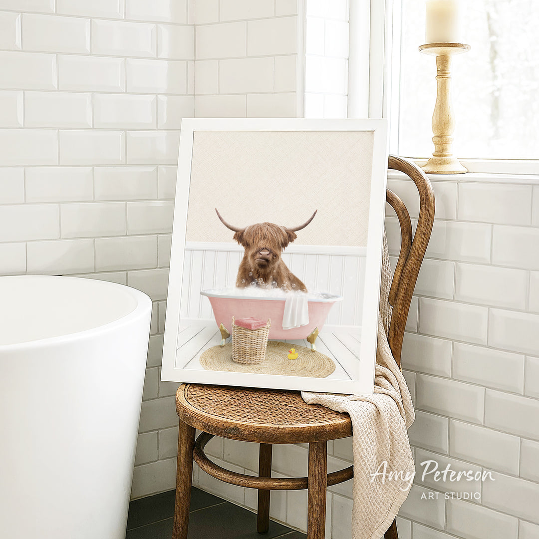 A framed image of a Highland cow sitting in a pink bathtub, with a towel and a basket nearby, is displayed on a wooden chair in a bathroom setting.