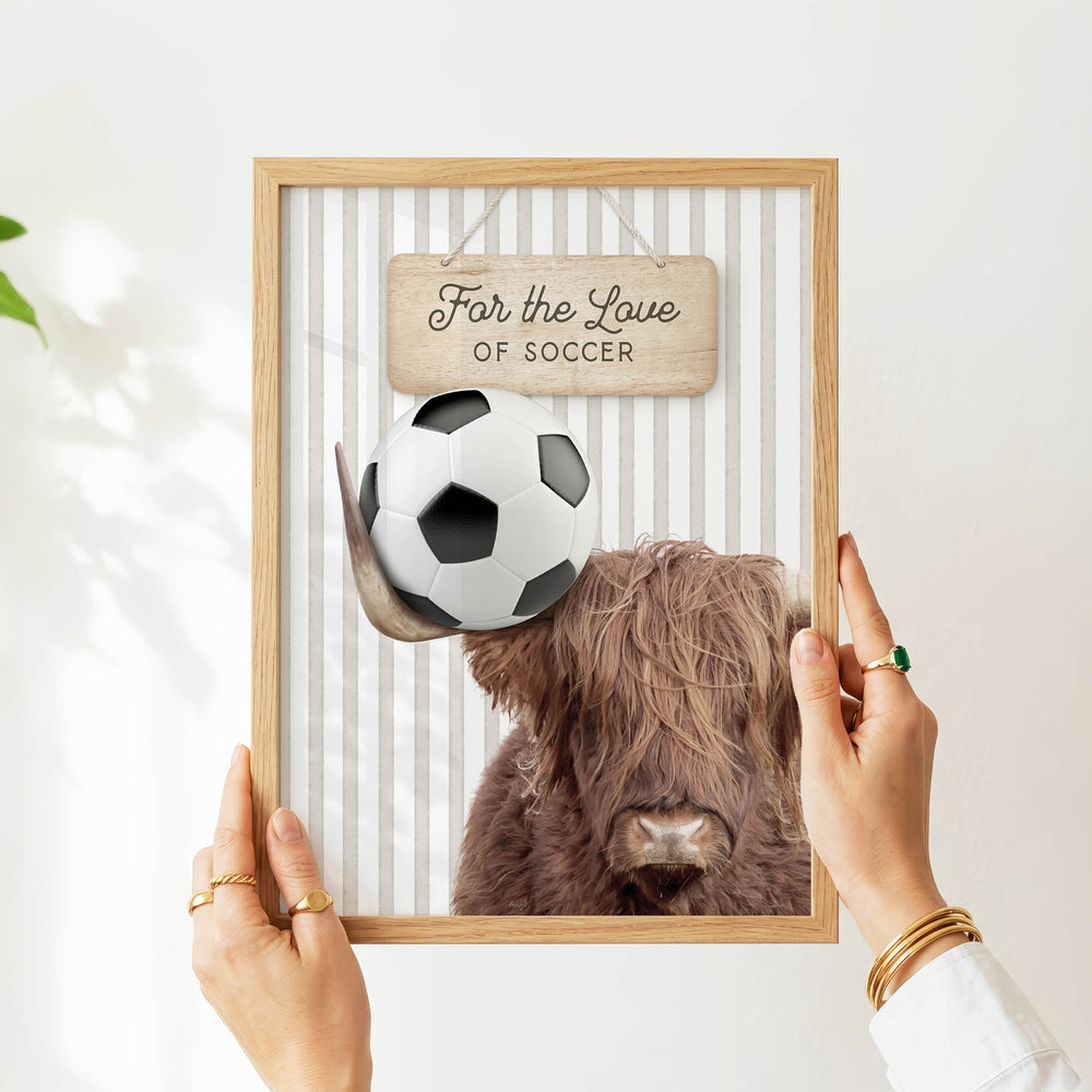 A person's hands are holding a framed picture of a Highland cow with a soccer ball above its head.