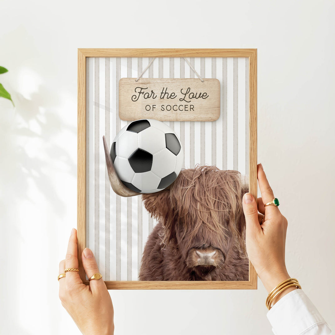 A person's hands are holding a framed picture of a Highland cow with a soccer ball above its head.