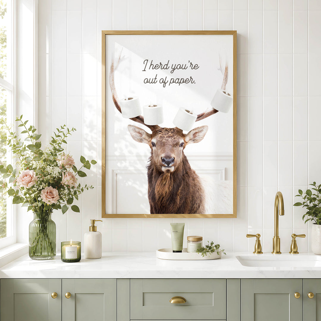a framed picture of a deer with antlers, surrounded by toilet paper, placed above a bathroom sink with a vase of flowers and other decorative items.