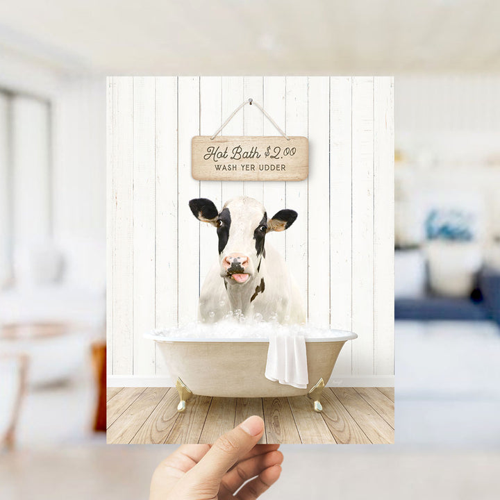 A hand holding a framed image of a cow sitting in a bathtub, with a sign above it that says "Hot Bath $2.00 WASH YER UDERDER".
