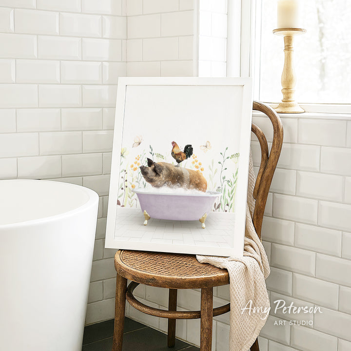 A framed illustration of a cat sitting in a bathtub, surrounded by flowers and a rooster, is displayed on a wooden chair in a bathroom setting.
