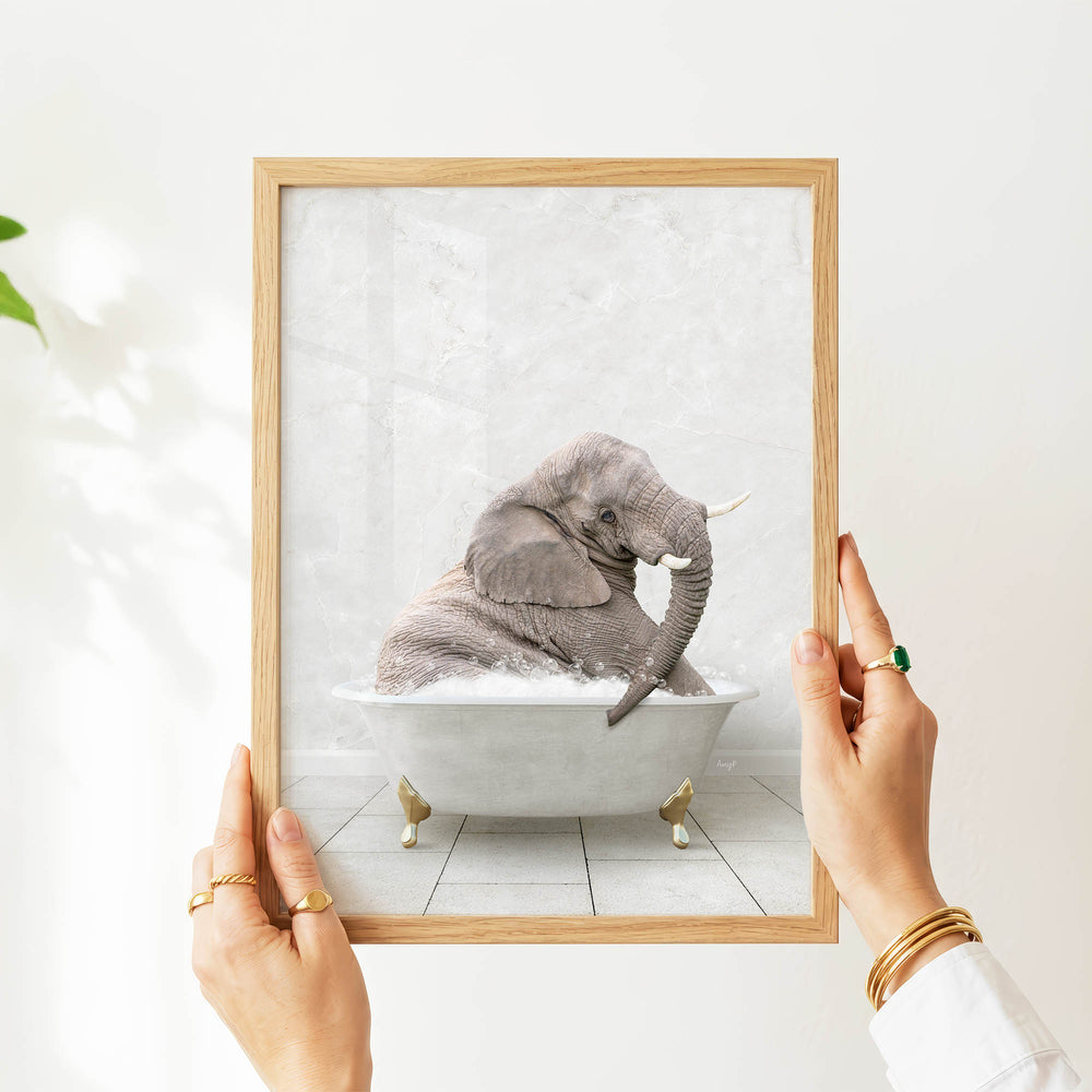A person's hands are holding a framed picture of an elephant sitting in a bathtub.
