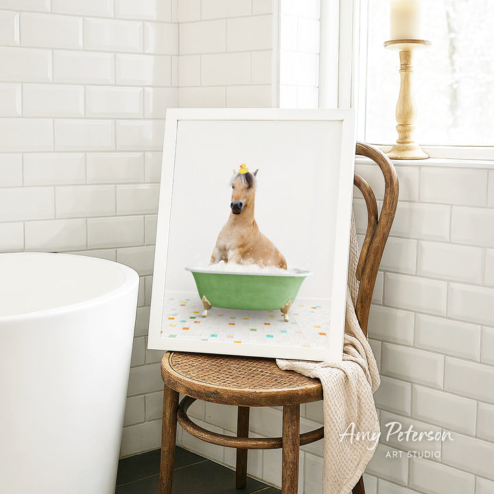 A framed image of a horse sitting in a green bathtub, surrounded by colorful tiles, is displayed on a wooden chair in a bathroom setting.