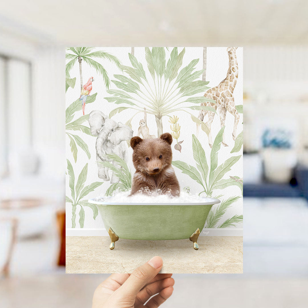 A hand holds up a framed print featuring a small brown bear sitting in a green bathtub, surrounded by tropical foliage and animals.