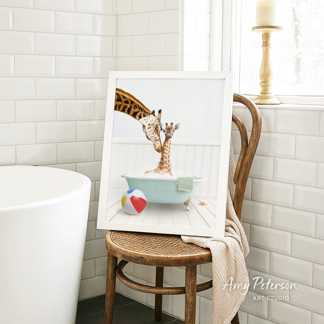 A framed image of two giraffes standing in a bathtub, with a beach ball and towel nearby, is displayed on a wooden chair in a bathroom setting.