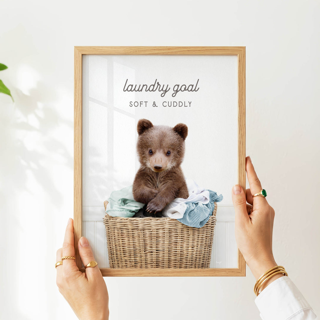 A person's hands are holding a picture frame with a picture of a baby bear sitting in a wicker basket filled with clothes.