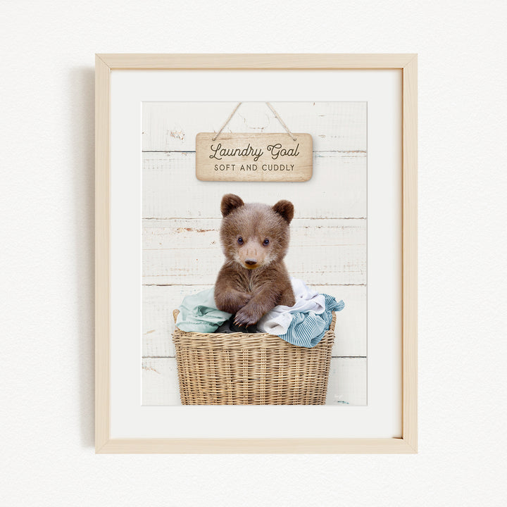 A framed image of a baby bear sitting in a wicker basket filled with clothes, with a wooden sign above that reads "Laundry goal Soft and cuddly".